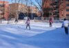 Adults playing ice hockey on an outdoor rink in a city during winter.