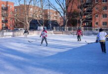 Adults playing ice hockey on an outdoor rink in a city during winter.