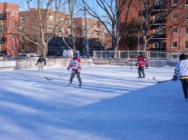 Adults playing ice hockey on an outdoor rink in a city during winter.