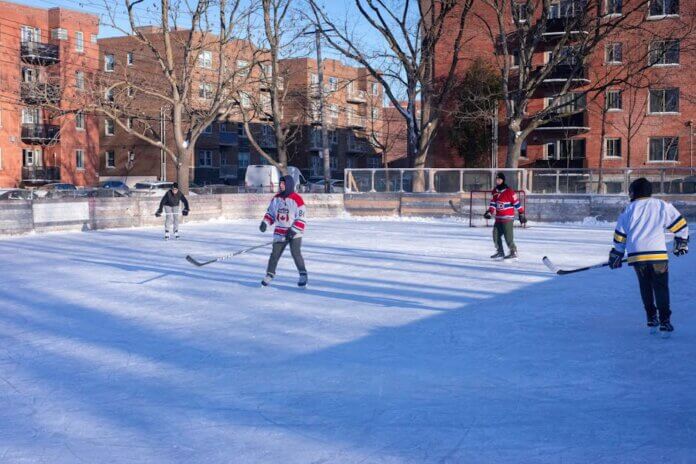 Adults playing ice hockey on an outdoor rink in a city during winter.