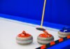 Curling stones with colorful handles and broom placed on ice sheet near blue wall and border during game in arena