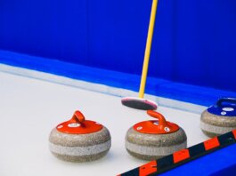 Curling stones with colorful handles and broom placed on ice sheet near blue wall and border during game in arena