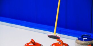 Curling stones with colorful handles and broom placed on ice sheet near blue wall and border during game in arena