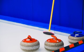 Curling stones with colorful handles and broom placed on ice sheet near blue wall and border during game in arena