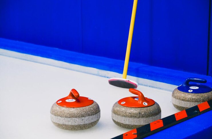 Curling stones with colorful handles and broom placed on ice sheet near blue wall and border during game in arena