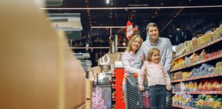 Family enjoying a shopping trip in a supermarket, with a child in a cart and parents smiling.