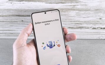 A close-up of a hand holding a smartphone displaying the One UI 6.1 welcome screen indoors.