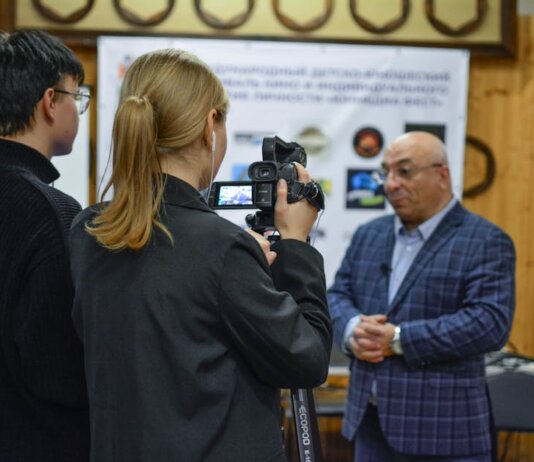 A journalist records an interview with a man using a camcorder indoors.