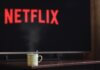 Консерваторы требуют отменить повышение «налога на Netflix» до 15% A steaming mug on a table in front of a TV displaying Netflix, creating a cozy indoor atmosphere.