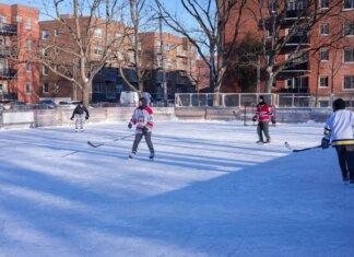 Adults playing ice hockey on an outdoor rink in a city during winter.