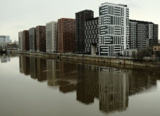 В Галифаксе подготовят кадры для строительства большего числа домов Captivating view of modern high-rise buildings reflecting in a calm river under cloudy skies.