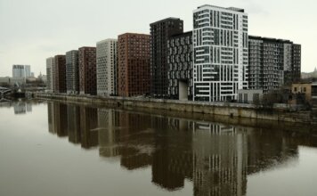 В Галифаксе подготовят кадры для строительства большего числа домов Captivating view of modern high-rise buildings reflecting in a calm river under cloudy skies.