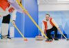 Determined sportsman throwing heavy granite curling stone near crop anonymous team sweeping ice sheet with brooms while playing sportive game