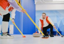 Determined sportsman throwing heavy granite curling stone near crop anonymous team sweeping ice sheet with brooms while playing sportive game