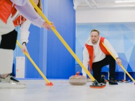 Determined sportsman throwing heavy granite curling stone near crop anonymous team sweeping ice sheet with brooms while playing sportive game