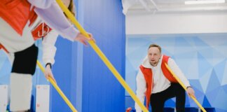 Determined sportsman throwing heavy granite curling stone near crop anonymous team sweeping ice sheet with brooms while playing sportive game