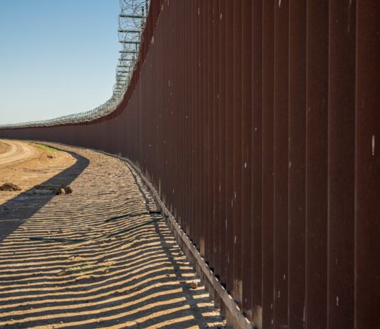 Long view of a steel border wall under clear skies, emphasizing solitude and division.