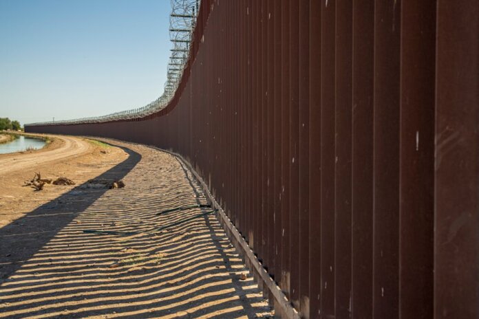 Long view of a steel border wall under clear skies, emphasizing solitude and division. Long view of a steel border wall under clear skies, emphasizing solitude and division.