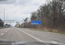 Vandalized road sign on a scenic highway surrounded by bare trees and cloudy sky.