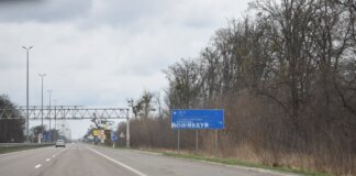 Vandalized road sign on a scenic highway surrounded by bare trees and cloudy sky.