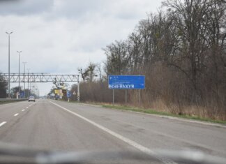 Vandalized road sign on a scenic highway surrounded by bare trees and cloudy sky.