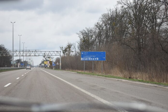 Vandalized road sign on a scenic highway surrounded by bare trees and cloudy sky.