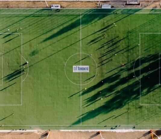 Aerial view of soccer field with anonymous sportspeople and shades against roadway on sunny day