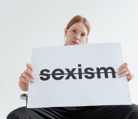 A caucasian woman confidently holds a sign addressing sexism in a bright studio setting.