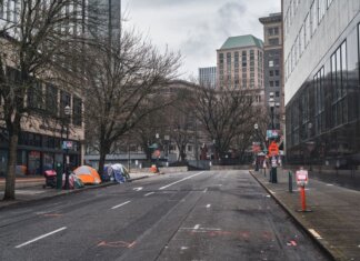 A city street with tents showing urban homelessness beneath bare trees on cloudy day.