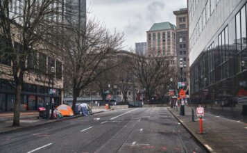 A city street with tents showing urban homelessness beneath bare trees on cloudy day.