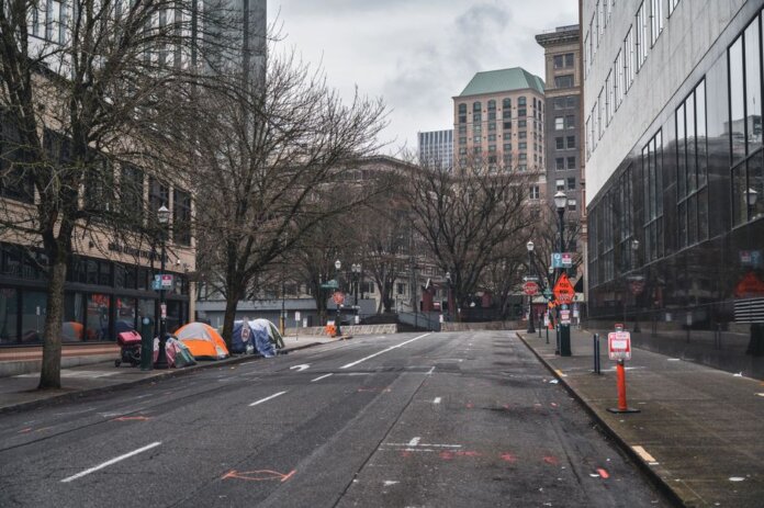 A city street with tents showing urban homelessness beneath bare trees on cloudy day. A city street with tents showing urban homelessness beneath bare trees on cloudy day.