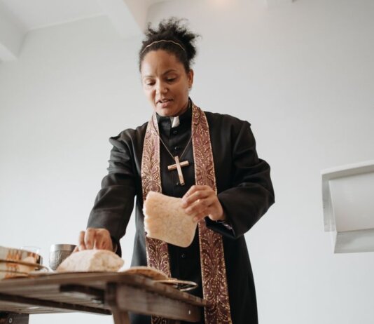 A female priest breaking bread at the altar during a religious service indoors, symbolizing unity and faith.