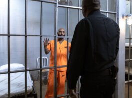 A male prisoner in an orange uniform stands behind bars facing a guard in a jail cell.