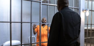 A male prisoner in an orange uniform stands behind bars facing a guard in a jail cell.