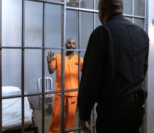 A male prisoner in an orange uniform stands behind bars facing a guard in a jail cell.