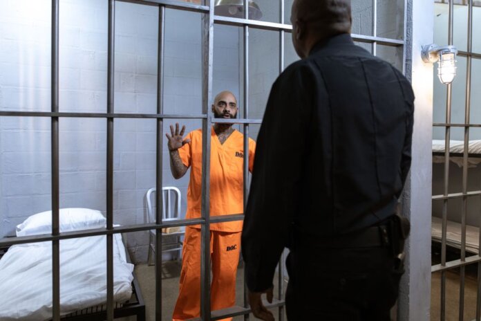 A male prisoner in an orange uniform stands behind bars facing a guard in a jail cell. A male prisoner in an orange uniform stands behind bars facing a guard in a jail cell.