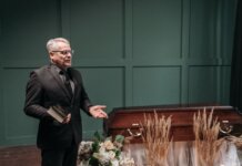 A solemn funeral ceremony featuring a pastor in a black suit delivering a speech next to a wooden coffin adorned with flowers.