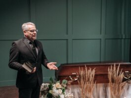 A solemn funeral ceremony featuring a pastor in a black suit delivering a speech next to a wooden coffin adorned with flowers.