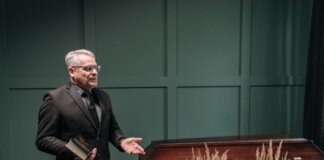 A solemn funeral ceremony featuring a pastor in a black suit delivering a speech next to a wooden coffin adorned with flowers.