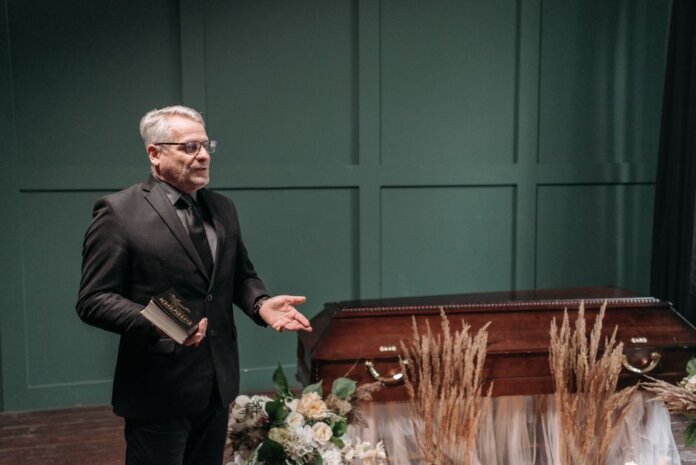 A solemn funeral ceremony featuring a pastor in a black suit delivering a speech next to a wooden coffin adorned with flowers. A solemn funeral ceremony featuring a pastor in a black suit delivering a speech next to a wooden coffin adorned with flowers.