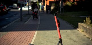 A Voi e-scooter parked on a sunlit city sidewalk with a blurred urban background.