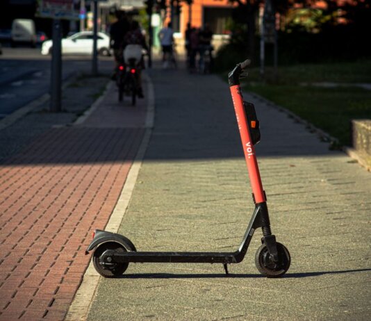 A Voi e-scooter parked on a sunlit city sidewalk with a blurred urban background.
