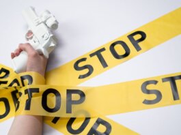 A white toy gun held by a hand wrapped in yellow stop tape on a white background, conveying a message of caution.