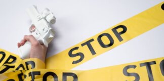 A white toy gun held by a hand wrapped in yellow stop tape on a white background, conveying a message of caution.
