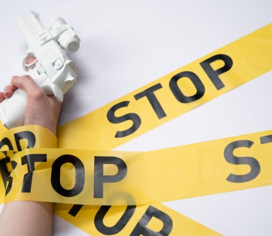 A white toy gun held by a hand wrapped in yellow stop tape on a white background, conveying a message of caution.