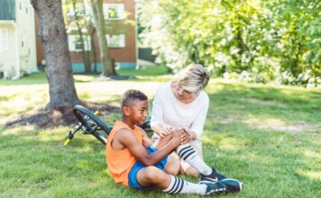 A woman comforts an injured child beside a bicycle in a sunny park setting.