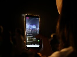 A woman live streams a concert using her smartphone at night. Vibrant stage in view.