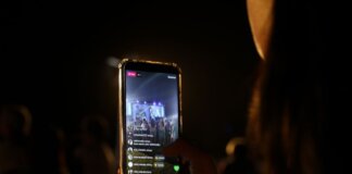 A woman live streams a concert using her smartphone at night. Vibrant stage in view.