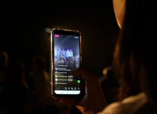 A woman live streams a concert using her smartphone at night. Vibrant stage in view.