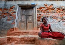A young girl sits calmly by a rustic brick wall with a wooden doorway, conveying resilience and tranquility.